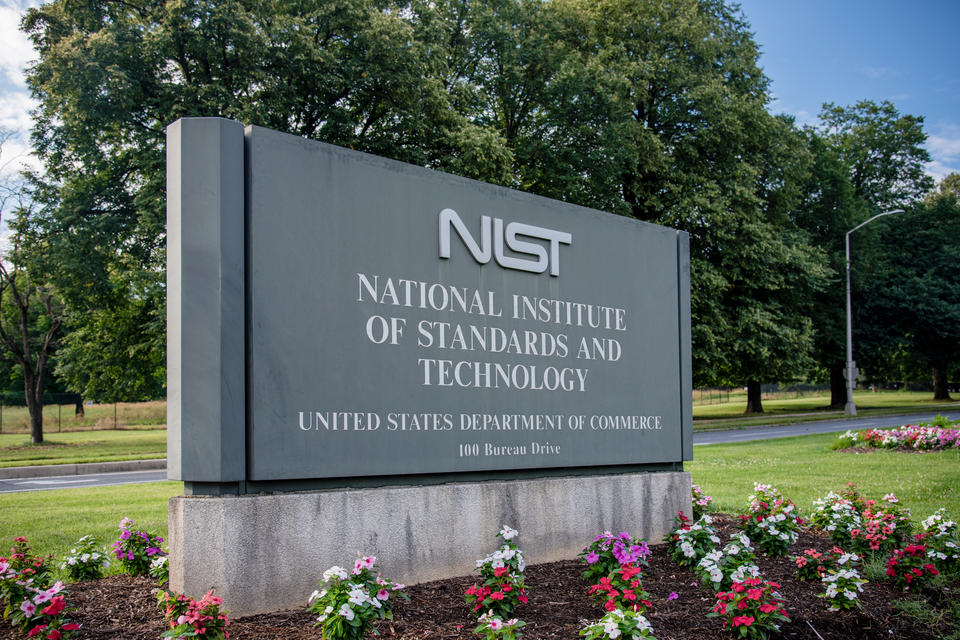 gray entrance sign that says NIST National Institute of Standards and Technology, United States Department of Commerce, 100 Bureau Drive. Surrounded by green grass, flowers, trees.
