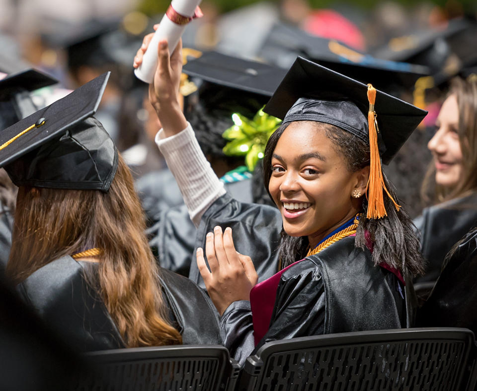 A female student at Howard Community College Commencement turning behind in her seat and smiling while holding up her diploma.