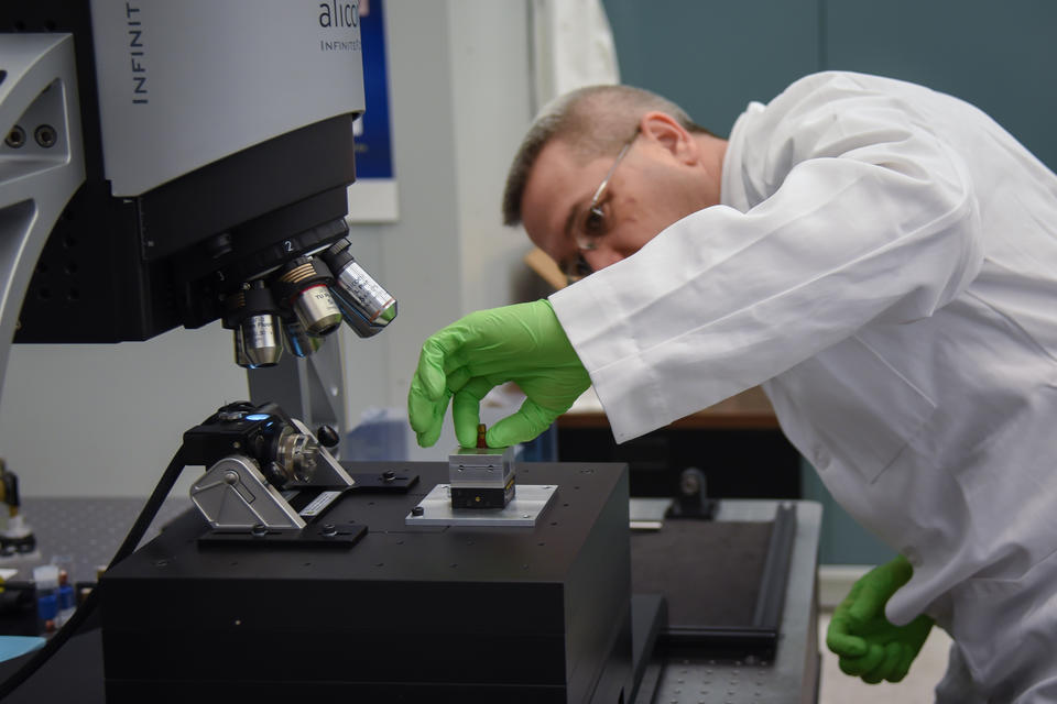 A man in a lab coat and green gloves places a bullet under a microscope.