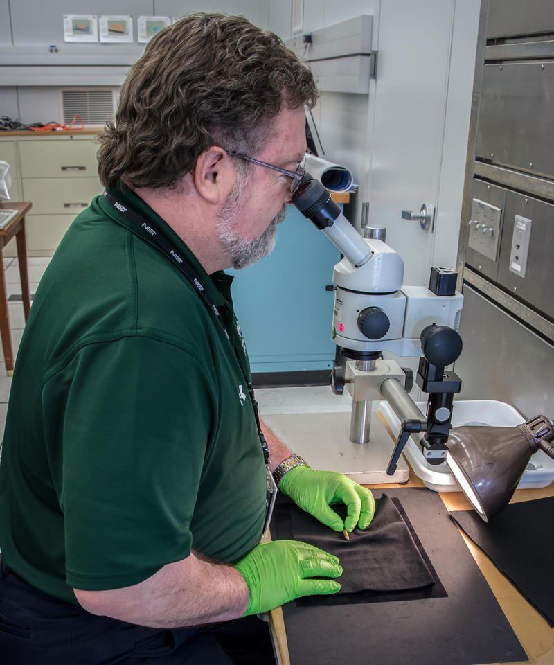 Man looks into microscope while holding bullet below.