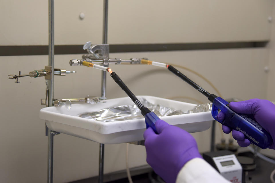 Two hands in purple gloves hold onto butane lighters, which are being used to light up two cigarettes poised in a metal device. 