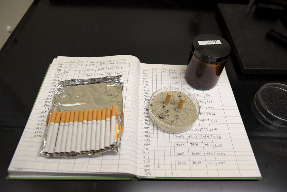 Several cigarettes, two extinguished butts, and a brown jar filled with cigarette smoke are sitting on top of a lab notebook full of notes. 