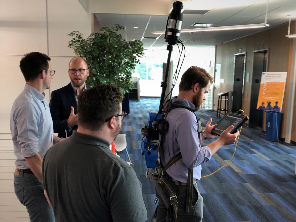 This is a photo of a LiDAR demonstration. A man is wear a backpack outfitted with LiDAR technology while three men speak around him.