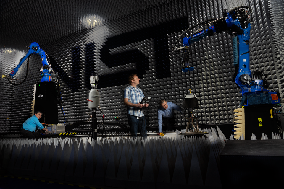 Researchers in large anechoic chamber with NIST logo on wall looking at robotic arms and equipment.