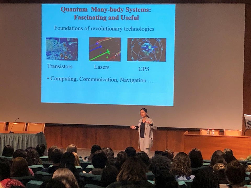 Woman on stage before a projected image that says "Quantum Many body systems: Fascinating and Useful. Foundations of revolutionary technologies: transistors, lasers, GPS. Computing, navigation, communication ...