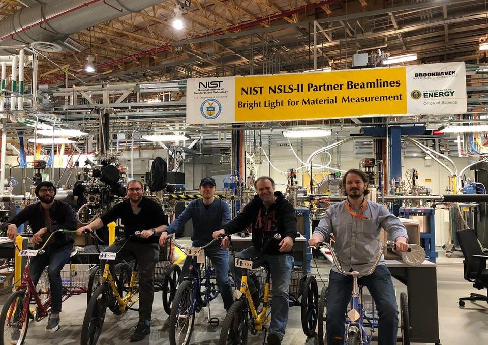 group of men parked on their bikes and smiling for the camera in a large guidehall. The sign hanging from the ceiling behind them reads "NIST NSLS-II Partner Beamlines, Bright light for material measurement"