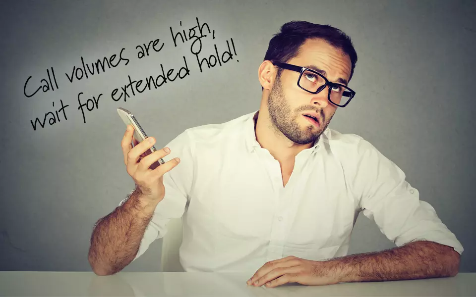 Frustrated man holding a cell phone listening to automated message saying "Call volumes are high, wait for extended hold!"