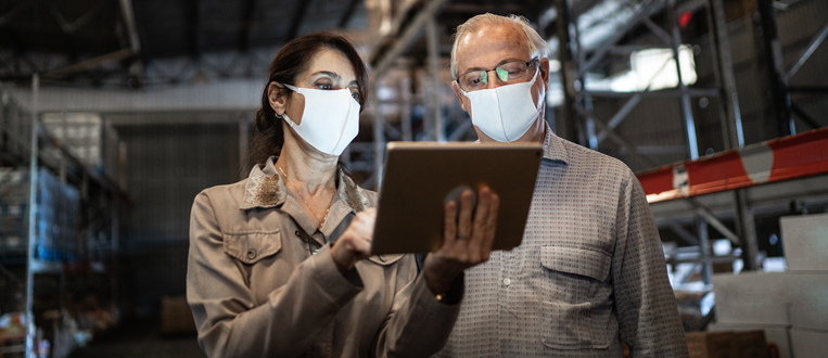 Senior partners walking and using digital tablet at warehouse with face masks