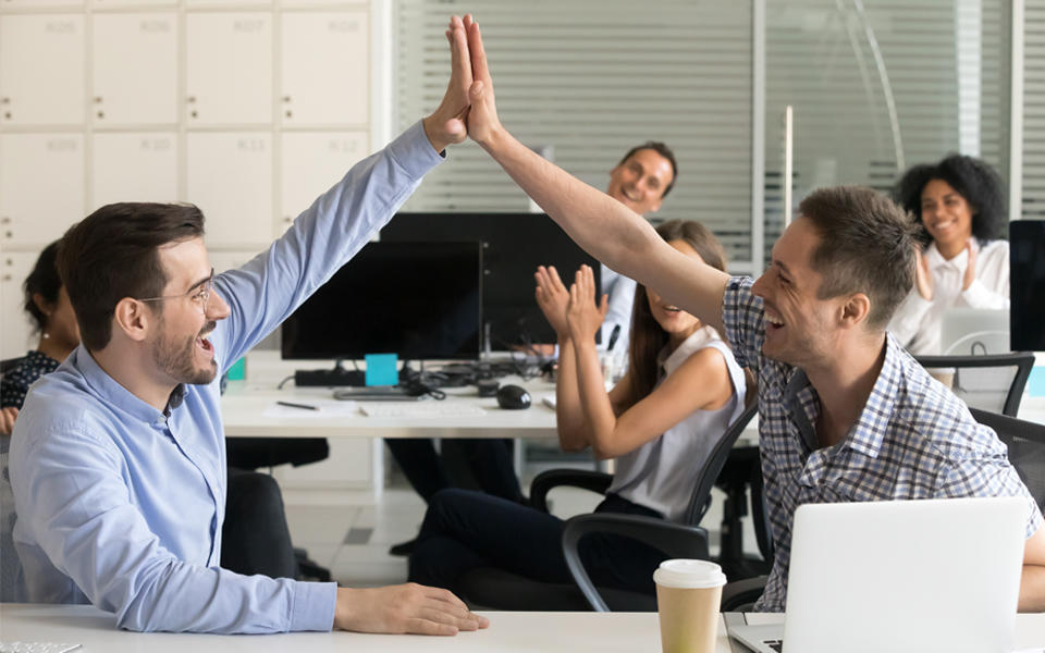Two male co-workers giving a high five with other female and male colleagues cheering them on.