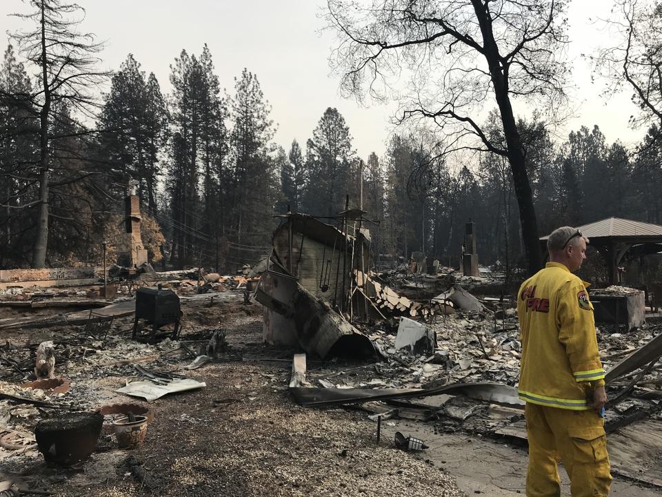 A man in a yellow fire jacket stands near burned buildings.