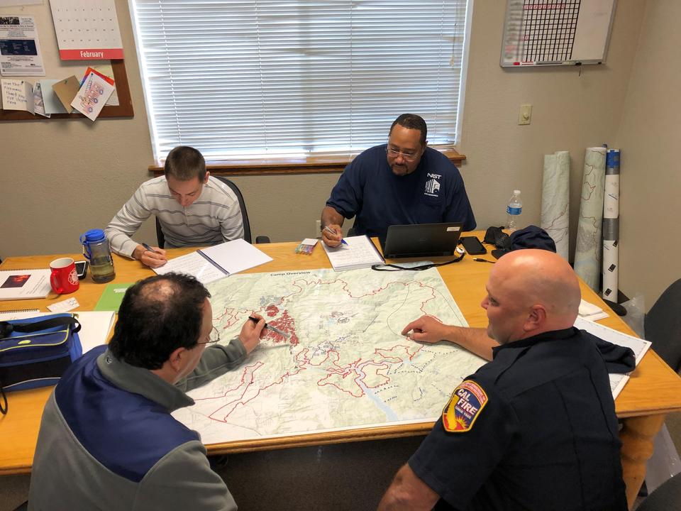 Four men sit around a table with a map spread on it.