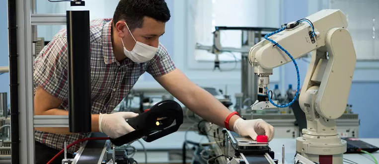 Man with surgical mask and gloves against COVID-19 ( Coronavirus ) programming robot arm with teach pendant.