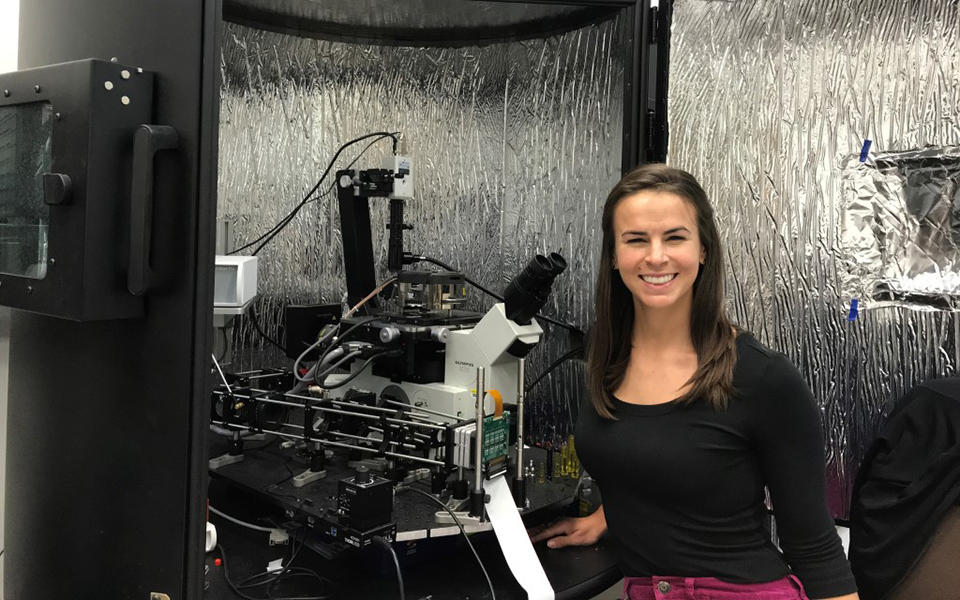 Callie Higgins in a lab. She is standing in front of a reflective screen surrounding an atomic force microscope. 