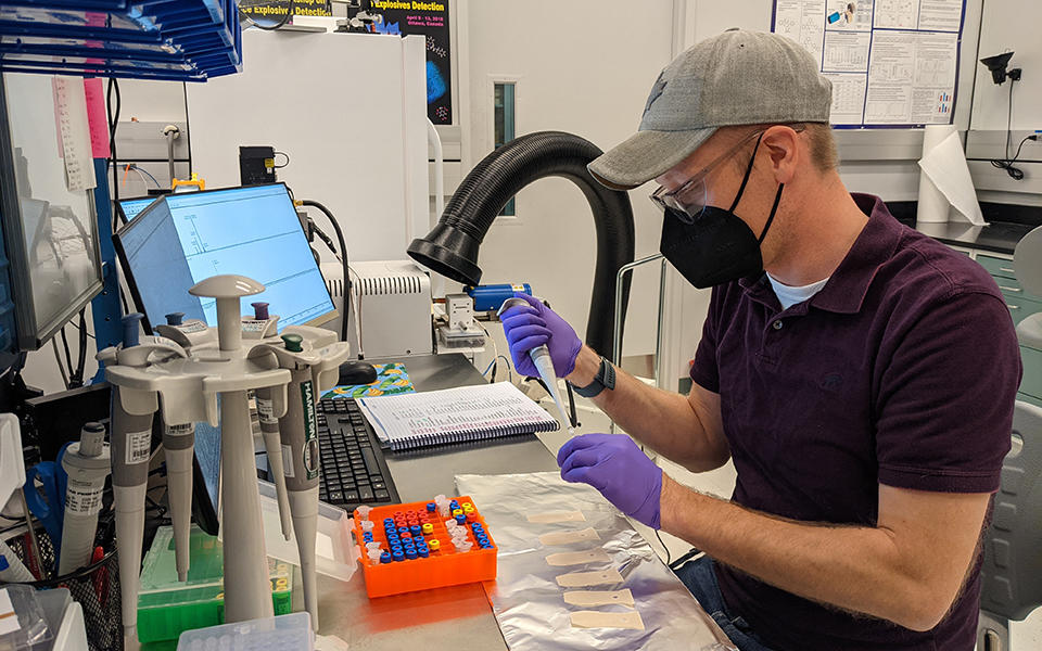 masked man with baseball cap seated at a lab bench pipetting samples 