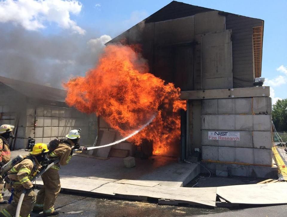 Firefighters spray water into a burning concrete building with a sign saying "NIST Fire Research."