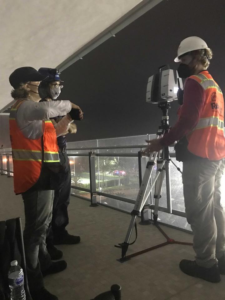 Three people in orange vests stand on a darkened balcony near a tripod.