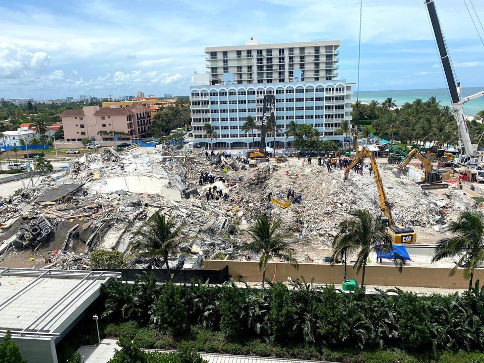 Palm trees stand in the foreground before a pile of debris with construction vehicles.