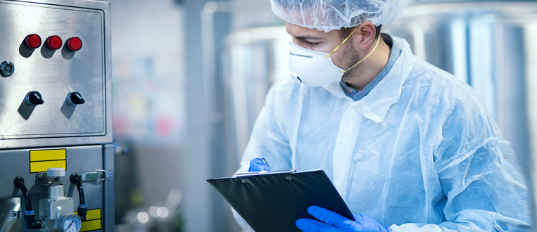 Technologist expert in protective uniform with hairnet and mask taking parameters from industrial machine in food production plant.