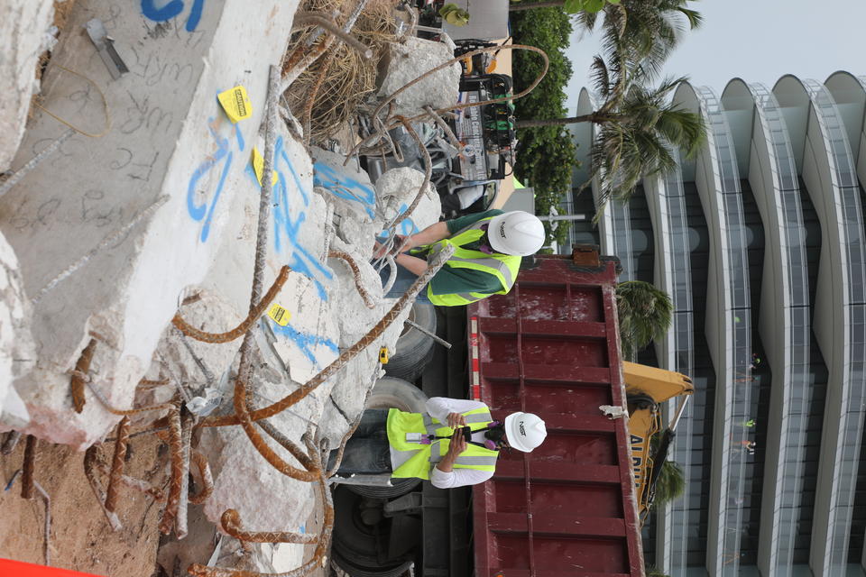 Two people in hard hats and other safety gear examine broken concrete columns with a dumpster and a condominium building in the background. 