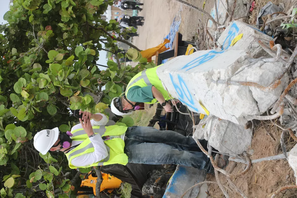 A man in a hard hat and other safety gear takes a picture of a broken concrete column with his phone.