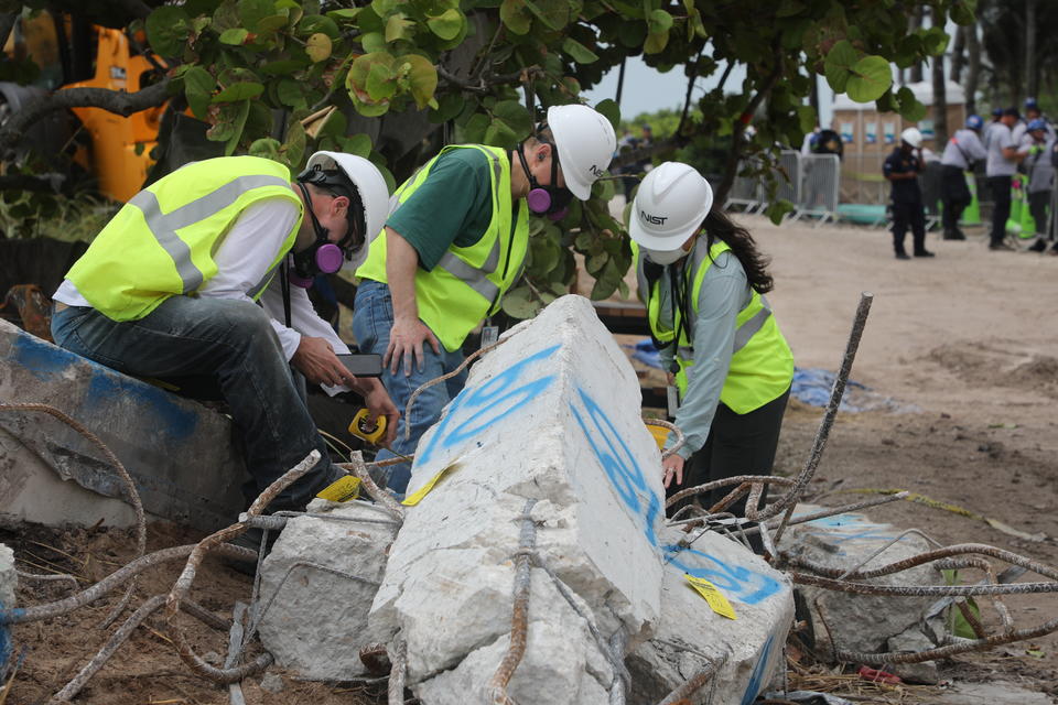 Three people in hard hats and other safety gear bend over a broken concrete column marked with blue spray paint. 