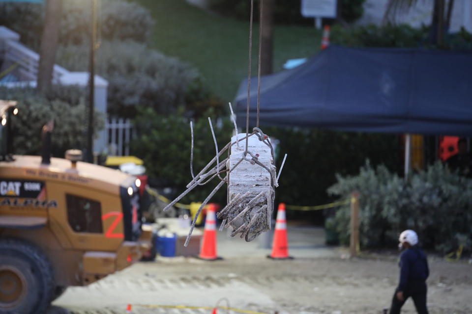 A piece of broken concrete column is lifted by a crane with construction equipment in the background.