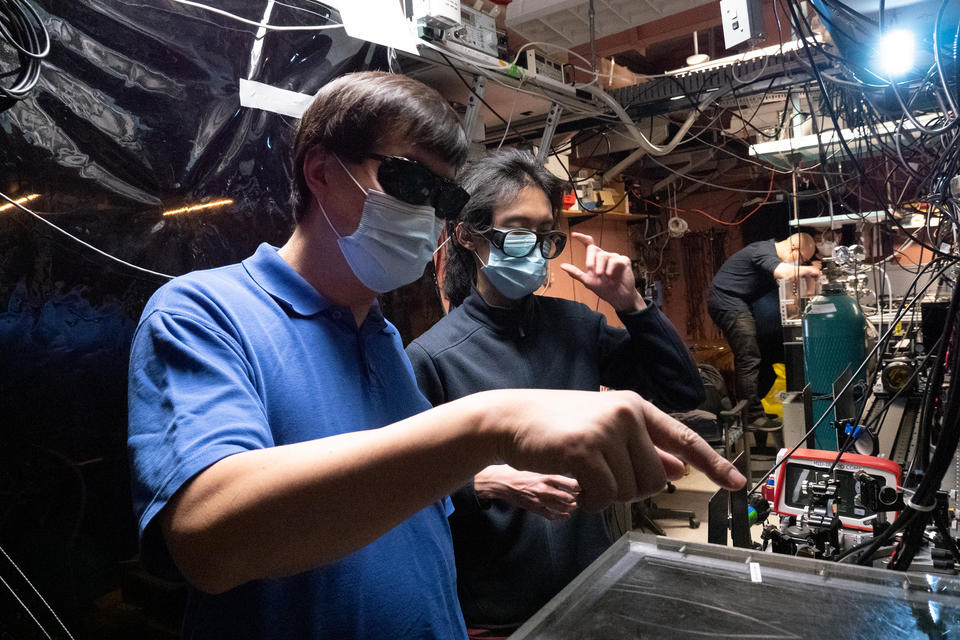 Jun Ye in mask on left pointing at a device. Man in mask stands beside him.