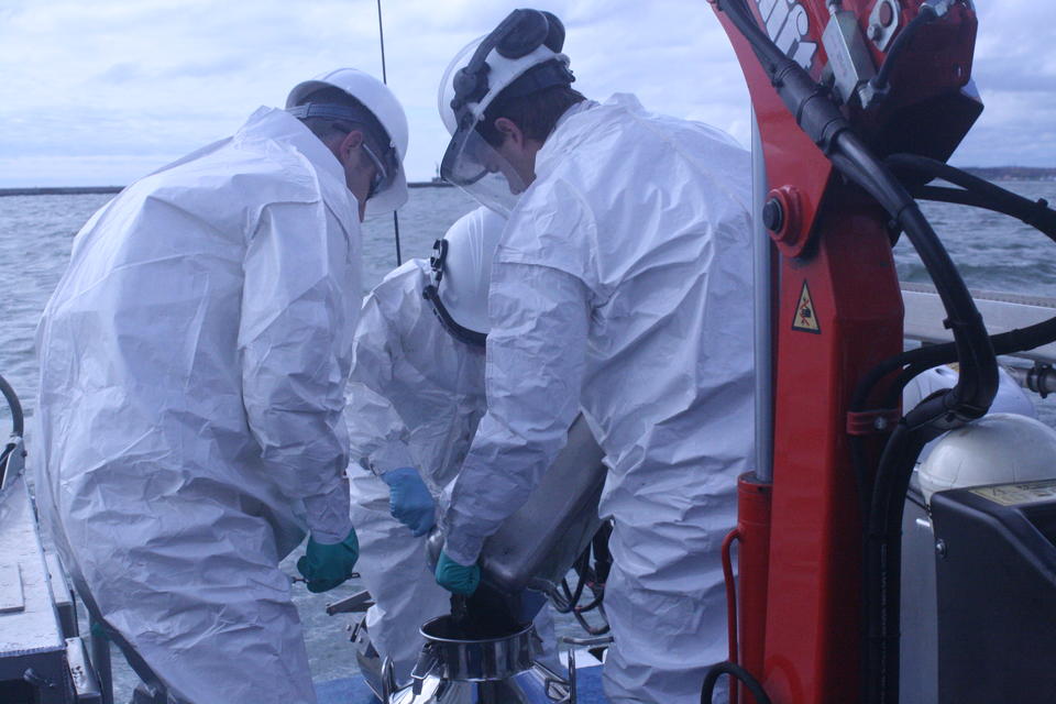 Three researchers on a boat wearing white overalls gather around to help pour material into a metal container.