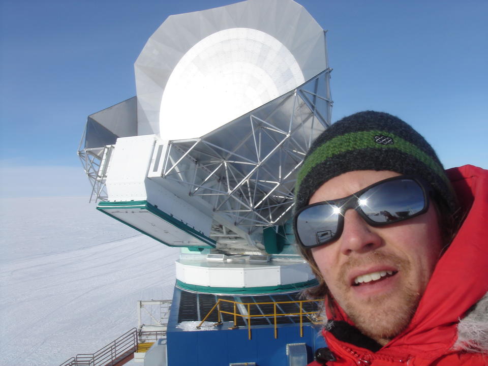 A man in winter gear stands in front of a large dish with a background of snow and blue sky. 