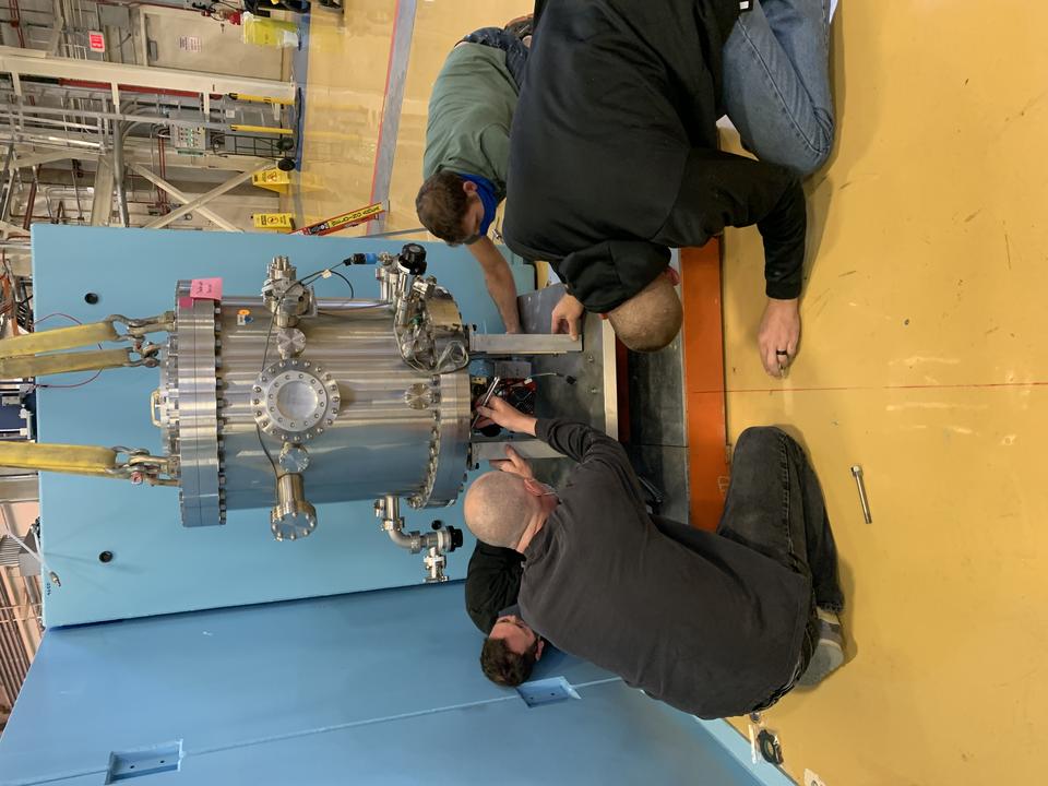 several men attach a hoisted aluminum chamber onto a mount below. 