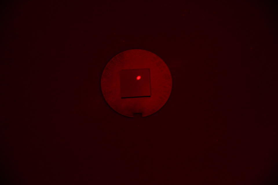 A square chip with a red laser dot lies on a round metal disk in a darkened space. 