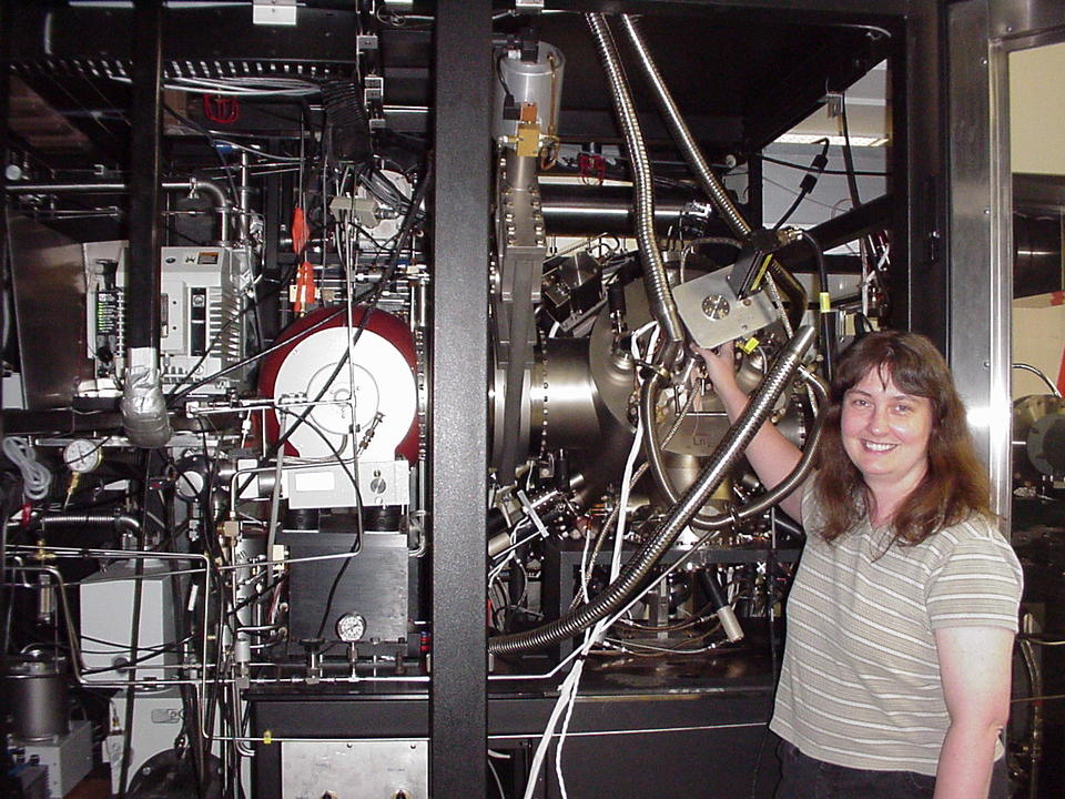 A woman stands smiling in front of a large device with many metal parts and wires.