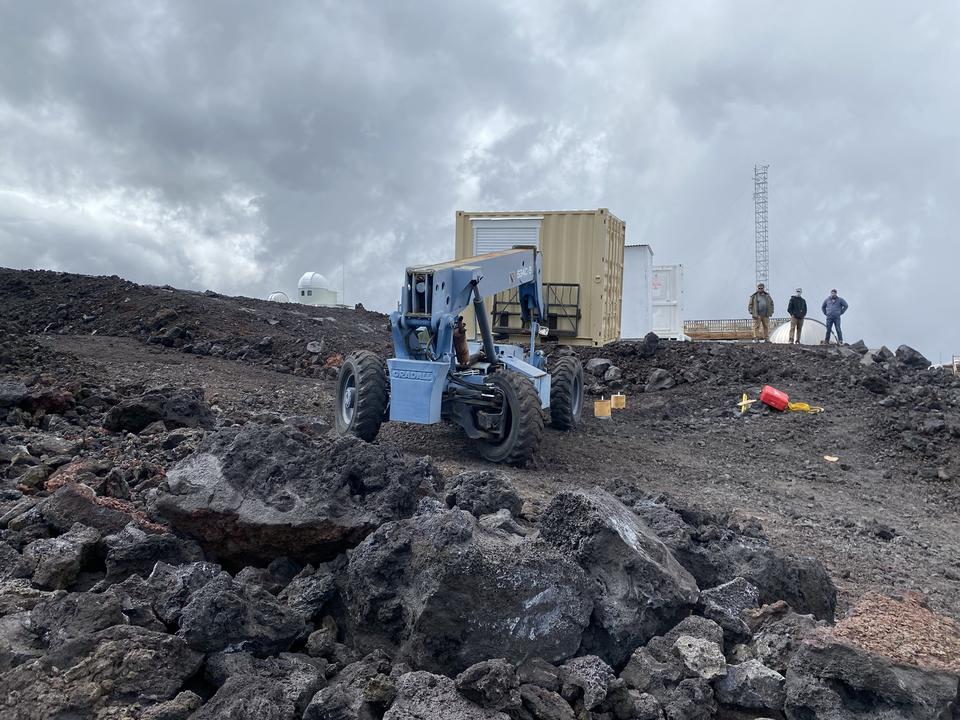 A blue construction vehicle sits near a pile of rubble with three people in the background.