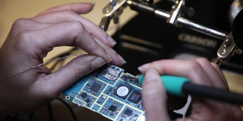 A closeup photograph of person's hands as they solder a wire onto a circuit board.