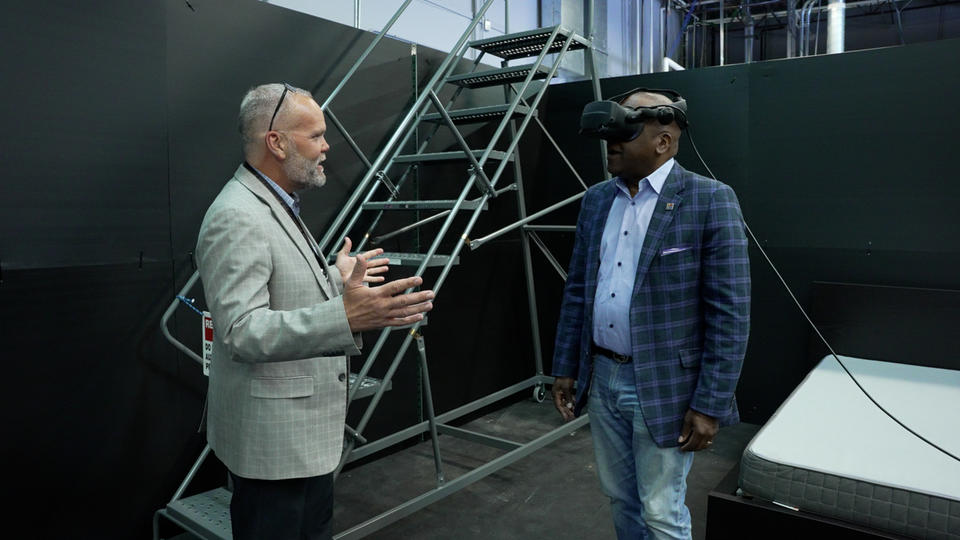 Two men, one wearing virtual reality goggles, standing in a virtual reality testing space