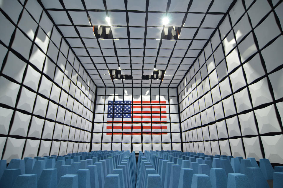 A room with grid-like walls of white boxes. Blue cone-shaped objects line the floor. An American flag is painted on the back wall.