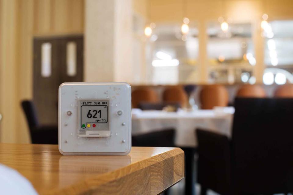 A white, square plastic CO2 meter sits on a table in a restaurant setting, reading 621 ppm.