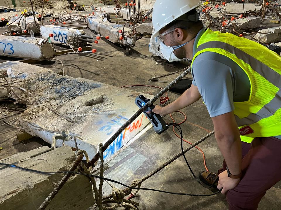 A researcher in safety gear leans over a large piece of concrete and rebar, pointing at it with a scanning device.