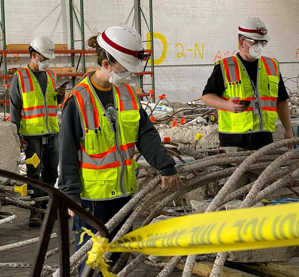 Three people in safety gear stand in a warehouse looking at chunks of concrete from Champlain Towers.