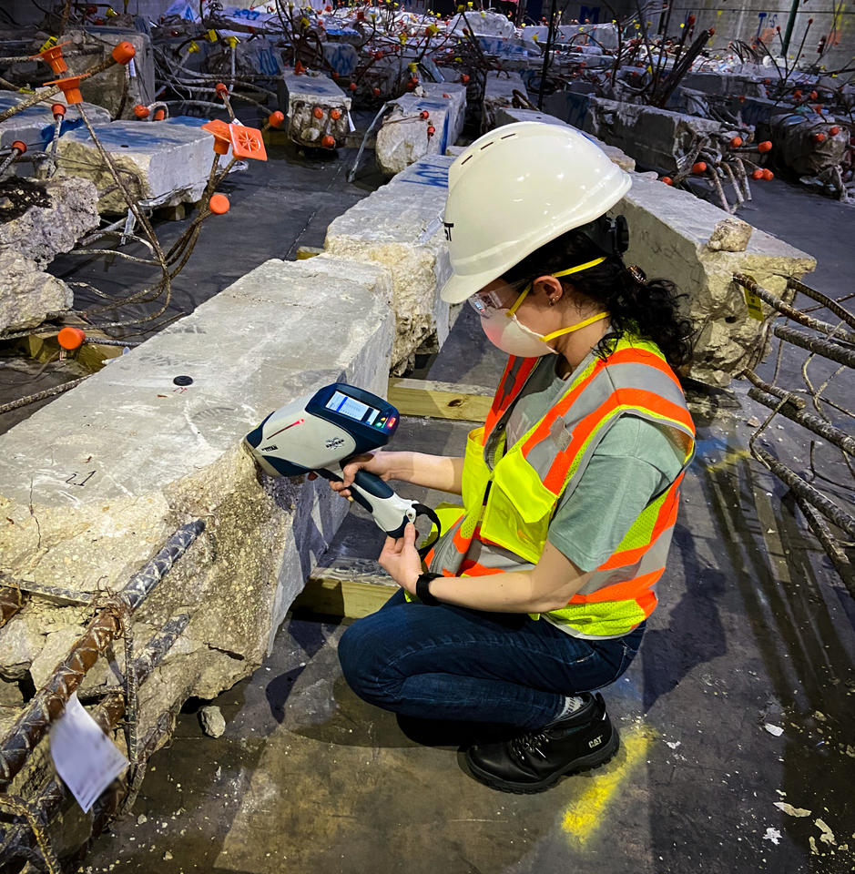 A researcher in safety gear crouches near a large piece of concrete, looking at a handheld device.