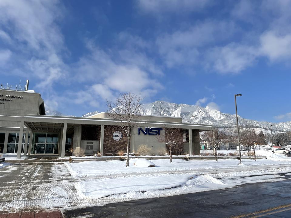 A low office building with a sign saying "NIST" is shown in front of mountains and a blue sky. Snow is on the ground.