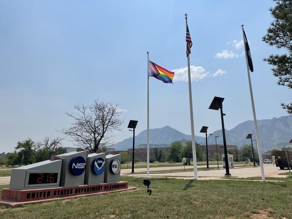 Entrance signs for NIST and NOAA are in the left foreground, with flagpoles, mountains and blue sky in the background.