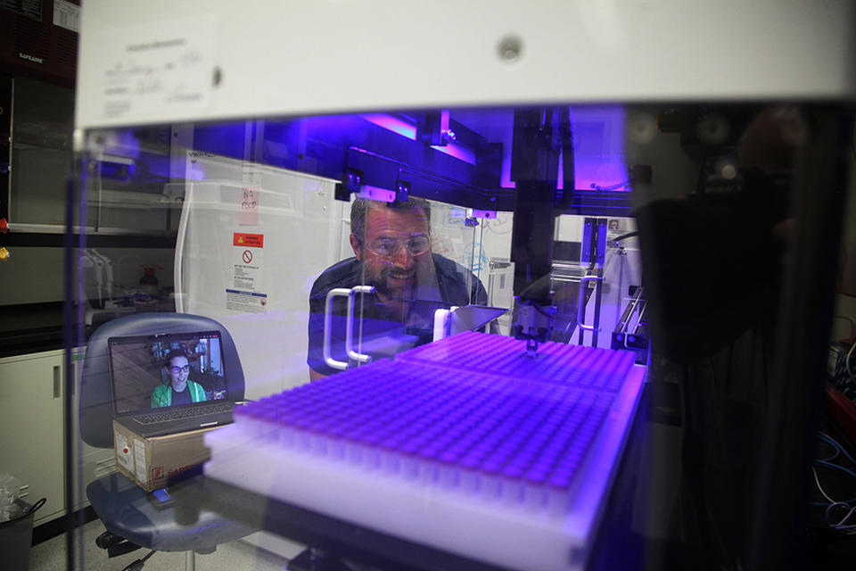 Two scientists watch as a robotic arms moves within a clear plastic box in a laboratory. The scientist on the left isn't there in person, but appears on a computer screen. 