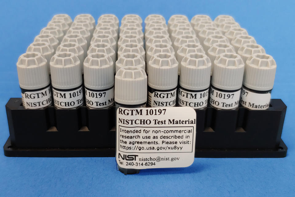 A rack of small vials with one pulled out in front for display, labeled "RGTM 10197 NISTCHO Test Material." 
