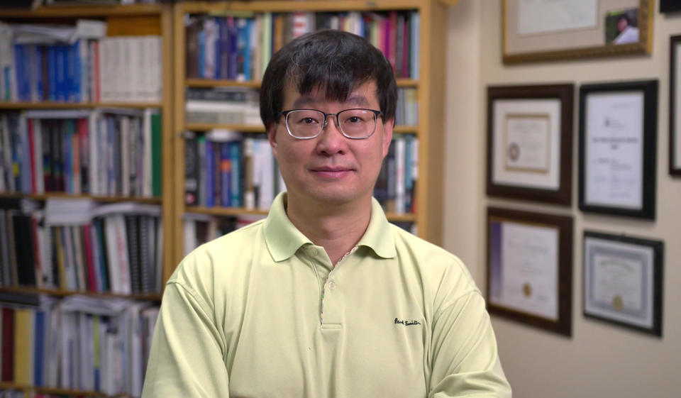 Jun Ye (from the chest up) standing in front of a wall of books and awards. Background is blurred out.