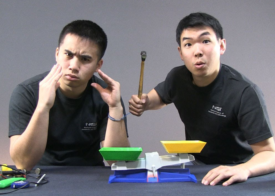 Screenshot from video shows two researchers in NIST T-shirts mugging over a plastic balance. 