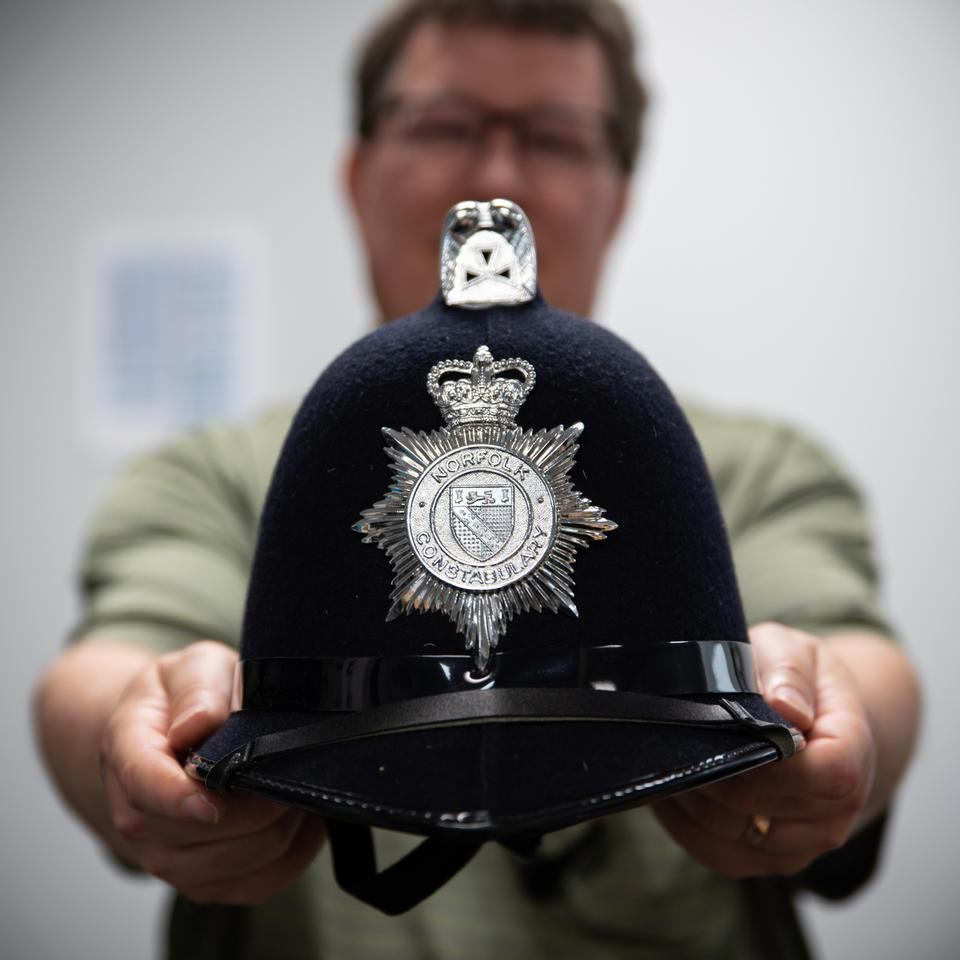 A blurry figure in the background holds out a black police helmet with a silver seal that reads "Norfolk Constabulary."