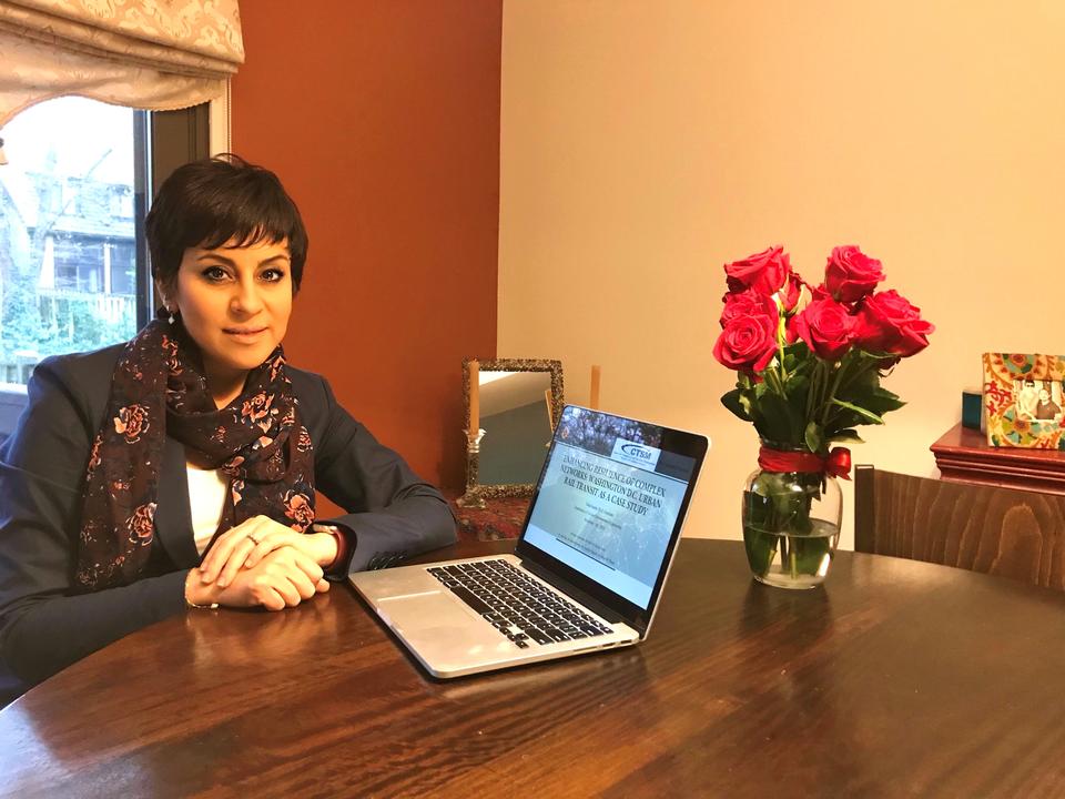 Yalda Saadat poses sitting at a table at home, next to an open laptop and a vase of roses. 