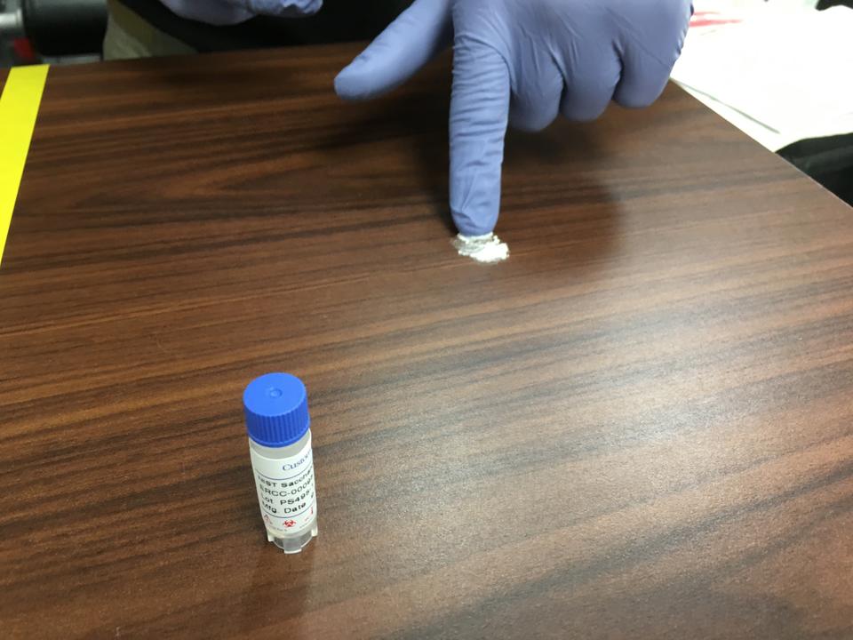 A person wearing purple plastic gloves points to a small amount of white powder on a tabletop. A plastic vial stands in the foreground.