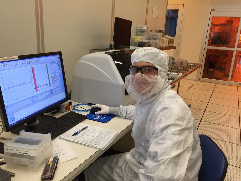 A person in a white coverall and safety glasses poses sitting at a computer in a lab.